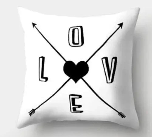 A white square throw pillow with the word LOVE" printed on it in black hollow letters. Each letter is on one side of the pillow. "L" s on the left, "O" is at the top, "V" is on the right, and "E" is on the bottom. There are two arrows forming an X crossing between the letters and a black heart in the center.