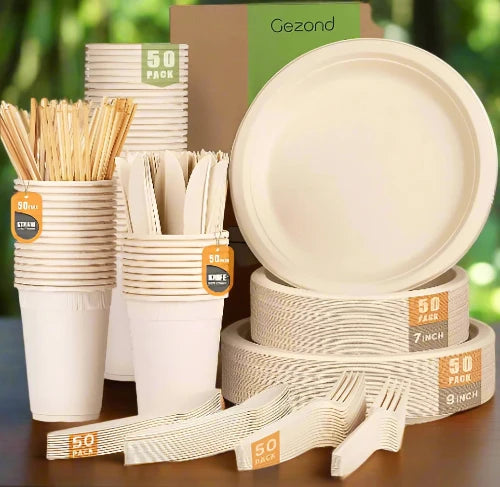 A set of cream-colored biodegradable disposable plates, cups and utensils are stacked, sitting on a wooden table to be used for a picnic.