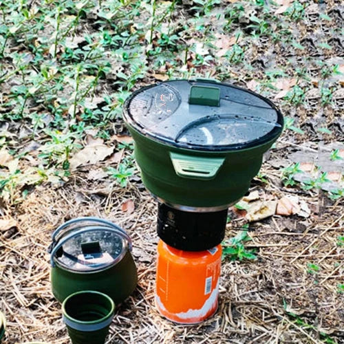 An army green collapsable silicone pot is atop an orange single burner stove at a campsite. It is surrounded by matching army green silicone collapsible cup and kettle with a lid.