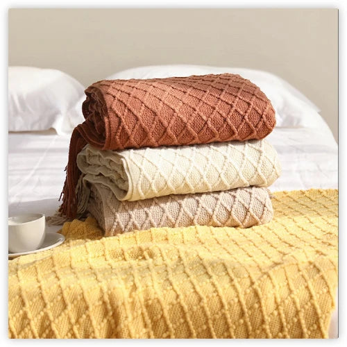 A stack of three knitted throw blankets in shades of brown, cream, and yellow resting on a bed. The blankets have a textured pattern and tassels at the ends.