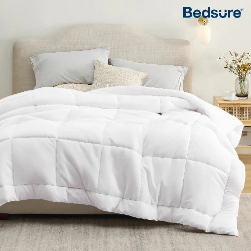 A cozy white duvet insert is imperfectly made on a bed of neutral tan and white colors with 3 pillows. A night table is on the left with a clear vase and flowers.