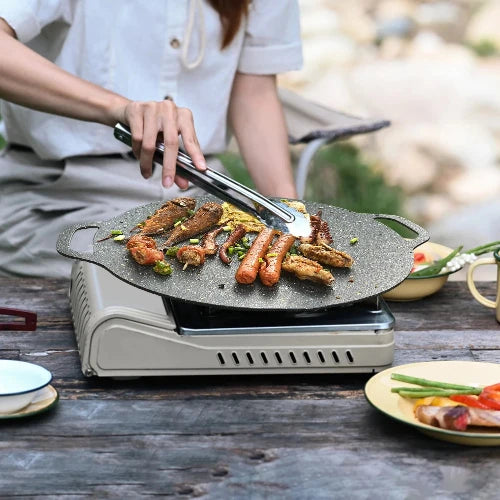 The black speckled griddle is being used to cook over a single burner stove outdoors on a wooden table with plates of food surrounding it.