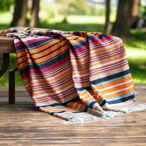 A brightly colored striped woven blanket is draped over the corner of a white bed. The blanket features stripes of blue, orange, pink, white and yellow.