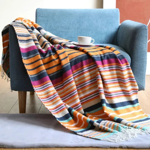 Family Glamping Striped Throw, colorful with orange, pink, cream, teal and navy stripe laid over a blue cushioned lounge chair with a mug and saucer resting atop the blanket.