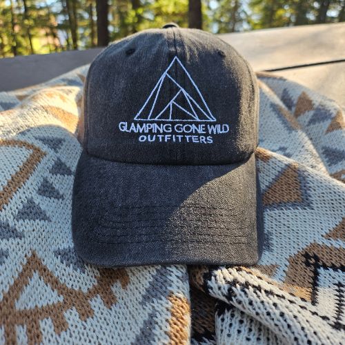 A black baseball cap with white embroidery that says, " Glamping Gone Wild Outfitters" is sitting on a blanket.