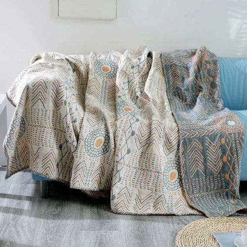 A cream-colored boho style throw blanket with various muted blue and orange geometric patterns is draped over the back of a light blue sofa. One side of the blanket is folded over showing the reverse side is muted blue with the designs in cream and muted orange.