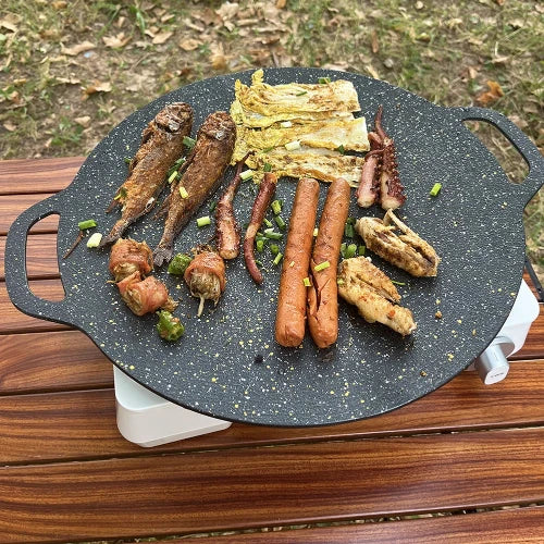 The black speckled griddle is shown featuring cooked meat and vegetables atop a wooden table.