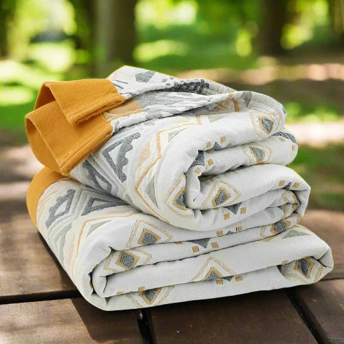 A white cotton gauze blanket with blue, grey, and yellow geometric shapes and a yellow border is folded and placed on top of a picnic table.