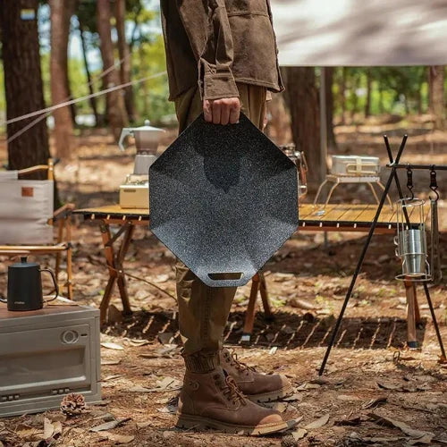 A person holding a griddle that has handles with one hand at their side while standing in a rustic setting.