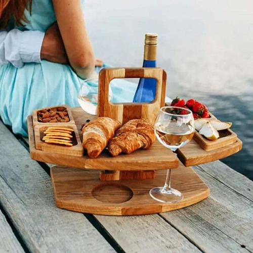 A wooden portable picnic table/tray designed for wine and snacks is sitting on a wooden table. The picnic tray has crackers, nuts, berries and apples in its snack holders, two croissants and wine bottle in a holder and a wine glass in a holder.