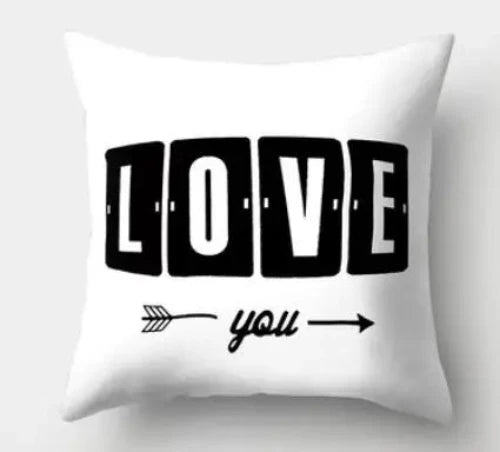 A white square throw pillow with the words, "LOVE you" on it. The word "LOVE" is in bold white font with each letter inside separate large rectangle black boxes. Under LOVE, an arrow points to the right under with the word "you" separating the front and back of the arrow