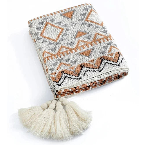 A cream colored knit blanket with grey, brown and black linear shapes is folded with all four of its tassels showing.