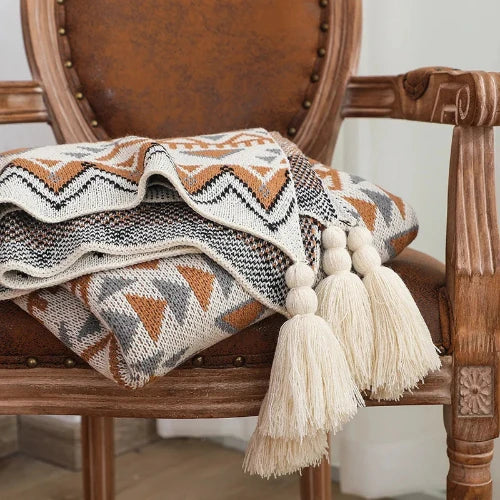 A knit blanket with a rustic motif in white, grey and brown is folded atop a wooden chair. The blanket has cream colored tassels at each corner.