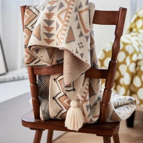 A cream colored knit blanket with angular shapes of grey, brown and black is draped over the back of a wooden chair. The blanket has tassels at each of its corners.