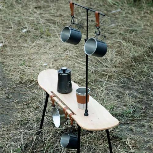 A brown wooden skateboard with black legs to make it a table has cups hanging from its side as well as from its metal T stand. A Kettle and coffee cup rest on the surface of the table in an outdoor setting.