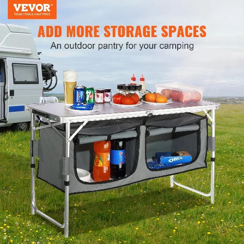 A camping table that offers more storage space for anywhere adventure awaits. It is set up outdoors with a camper van in the background. The table has two large storage compartments underneath. It is covered in various camping supplies, including drinks, food, and condiments. Text on the image reads: "VEVOR: ADD MORE STORAGE SPACES. An outdoor pantry for your camping."