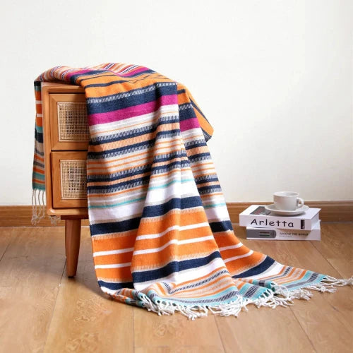 A brightly colored striped woven blanket is draped over the side of a small wooden nightstand. The blanket has stripes of orange, blue, pink, white, and green. The floor is hardwood and there is a white cup sitting on two books next to the nightstand.