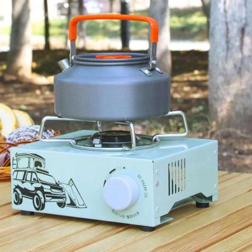 A light teal single burner butane gas stove with a black stenciled SUV camper and a temperature dial. The stove has a kettle on top and is in an outdoor camping site.