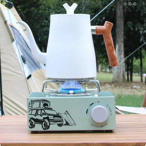 A light teal singe burner butane gas stove with a black stenciled SUV camper and a temperature dial. The stove has a white kettle on top and is in an outdoor camping site.