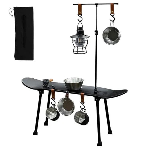 A black skateboard with four legs attached to make it a table has two cups and a bowl hanging from its side and a lantern and pot hanging from its T stand. There is a bowl resting on top of the table. In the top left area of this picture there is a carrying case.