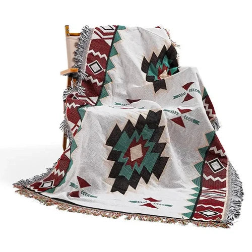 A cream colored blanket with geometric shapes creating a southwestern pattern in red orange, dark teal and black. All of the edges of the mat are tussled.