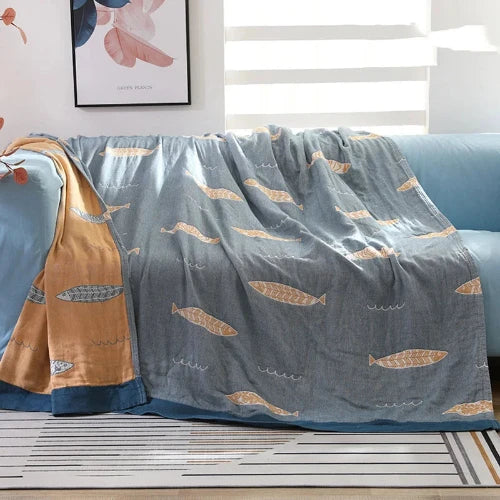 A reversable throw that is light blue with a repeating yellow fish design on one side and is yellow with a light blue repeating fish design on the other side. The blanket is spread out over a couch and one corner is folded to show its reversable design.