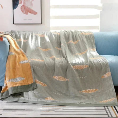 A reversable throw that is sage green with a repeating yellow fish design on one side and is yellow with a sage green repeating fish design on the other side. The blanket is spread out over a couch and one corner is folded to show its reversable design.